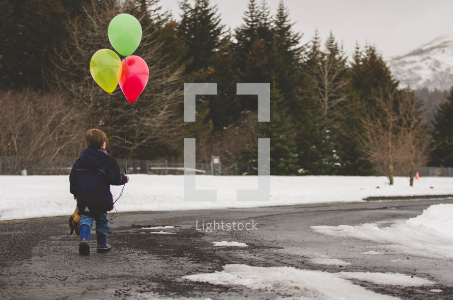 boy child walking in snow carrying a stuffed animal and balloons 