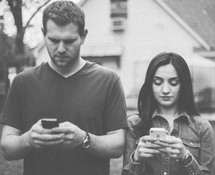 couple standing next to each other texting on a cellphone 