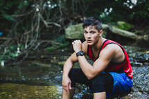 a young man sitting on a rock by a creek outdoors 