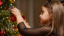 young girl putting a red ball to decorate Christmas tree 
