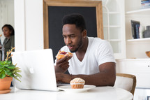 man eating a pastry while sitting in front of a laptop computer 