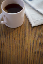 A cup of coffee next to a newspaper on a wooden table.