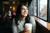 a smiling woman sitting in a window seat in a coffee shop holding a coffee 