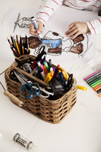 A child coloring at a table with a basket full of pencils and markers.