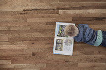 A toddler boy lying on a wood floor reading a book. 
