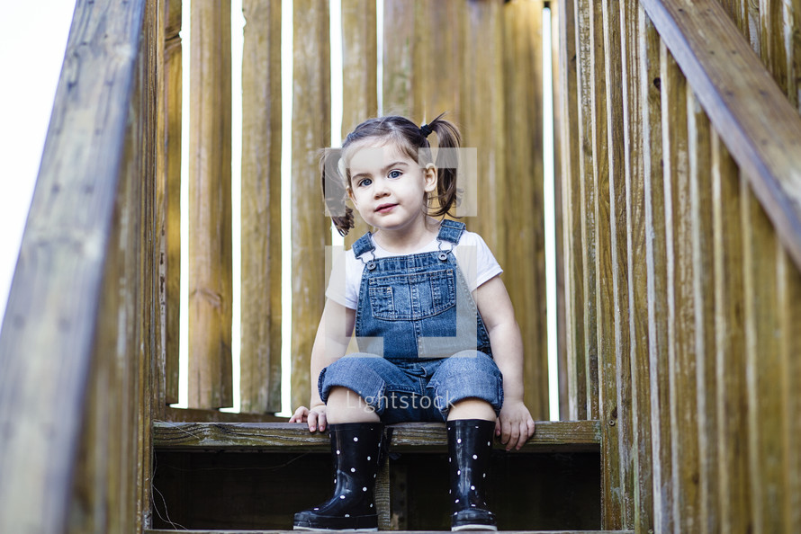 Small girl in overalls and boots, sitting on a wooden step 