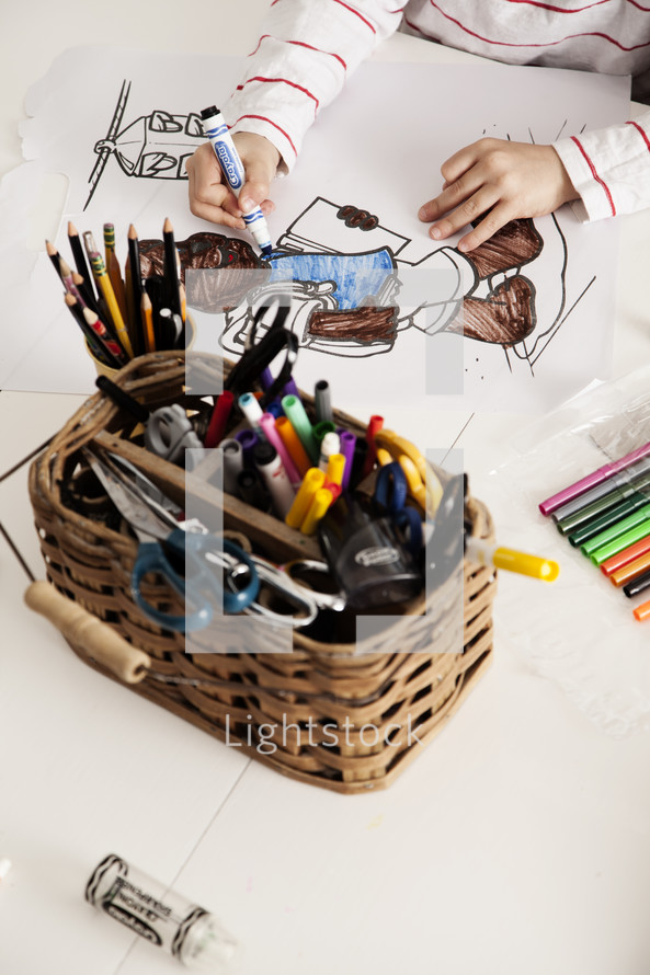 A child coloring at a table with a basket full of pencils and markers.