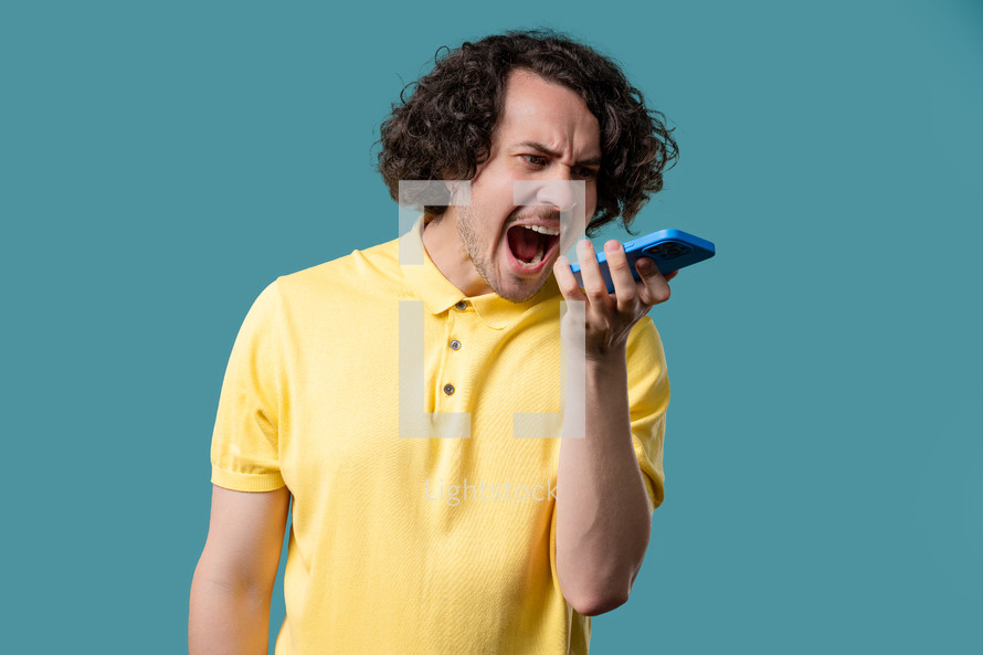 Annoyed young man quarreling while talking on mobile phone. Angry dude, stress. Blue studio background. Conflict, dispute, negative. High quality