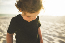 a toddler boy looking down at the sand on a beach 