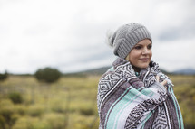 A woman wrapped in a blanket and wearing a winter hat stands near a field.