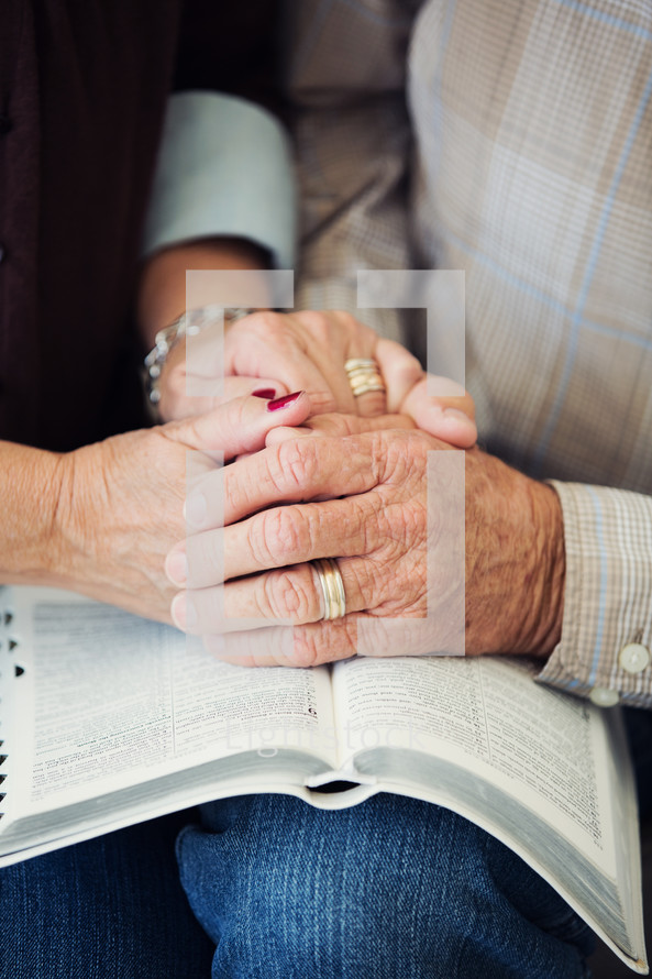 an elderly couple holding hands and praying over the pages of a Bible.