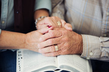 elderly couple holding hands and praying over the pages of a Bible 