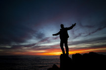 a man with outstretched arms standing on a rock by the ocean at sunset 