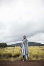 A woman wrapped in a blanket stands on a gravel road near a field.