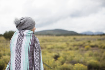 A woman wrapped in a blanket looks out over a field toward mountains.