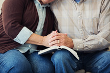 A mature couple praying together over the open pages of a Bible.