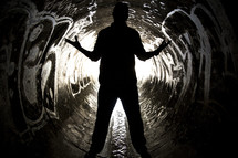 Silhouette of man straddling water flowing through a graffiti-painted sewer drain pipe with arms extended.