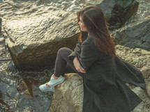 a young woman sitting on a rocky shore thinking 