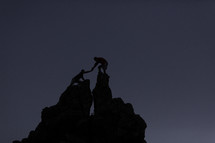 a man helping a woman reach the top of a rock peak 