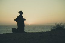 silhouette of a graduate on a beach kneeling in prayer