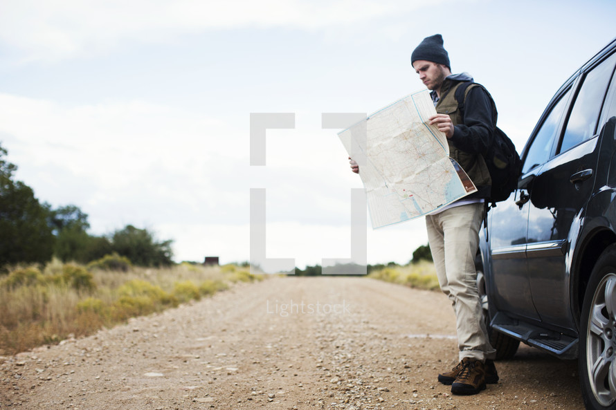 A man standing by his car on a gravel road while studying a road map.