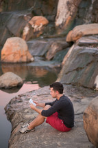 a man sitting on a rock shore of a river reading a Bible 