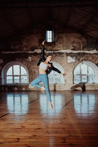 Young woman jumping in casual style - jeans and leather jacket doing ballet in old studio. Attractive ballerina practices in choreography alone. High quality photo