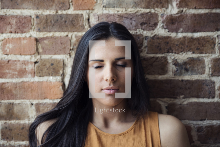 A young woman sitting against a brick wall with her eyes closed.