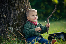 Little Cute Toddler with Wooden Stick Sitting by the Tree