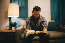 A man reading a Bible while sitting on a couch in his living room. 