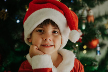 Portrait of adorable boy in Santa Claus costume on Christmas tree background. Home celebration, decoration with lights. Happy childhood, kids, lovely son. High quality photo