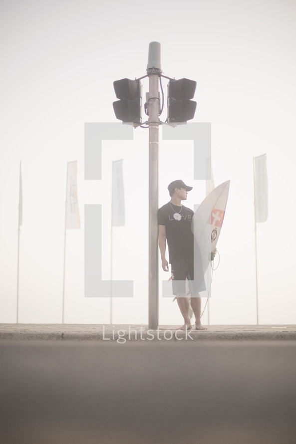 Man with surfboard leaning against a speaker pole on the beach.