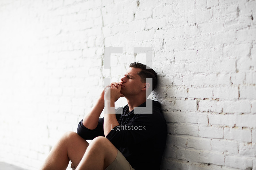 a man sitting alone leaning against a wall praying 