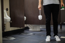 man in a public restroom holding a toilet brush 
