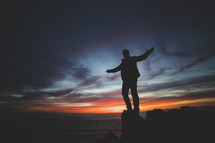 a man standing with outstretched arms on a shore at sunset 