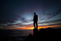 man standing on a rock along a shore at sunset 