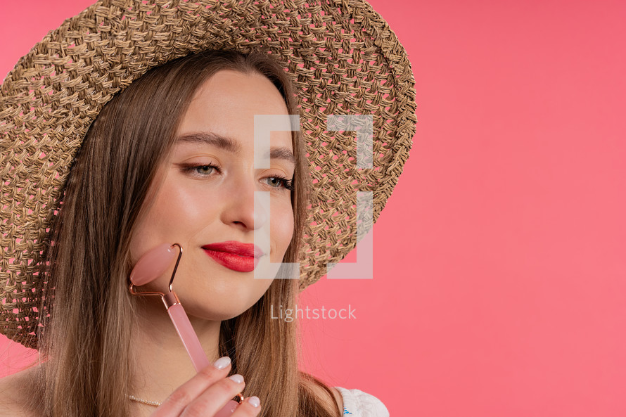 Young woman with rose quartz stone roller on pink background. Facial self care, beauty rituals, cosmetology, anti aging and anti-wrinkle treatment. Copy space. High quality photo