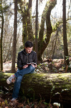 Man sitting on a fallen tree in the woods reading his Bible.