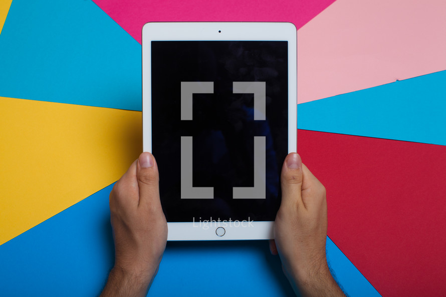 Hands holding an electronic tablet on a multi-color background.