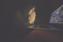 A man in a hooded sweatshirt praying in front of a computer screen.