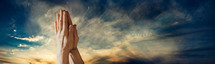 Hands praying against a sunset sky