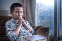 a boy praying in a window with a Bible in his lap 