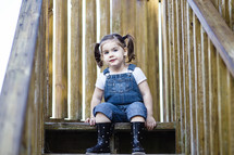 Small girl in overalls and boots, sitting on a wooden step 