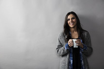 a young woman holding a coffee mug and smiling against a blank wall.