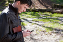 Man standing outdoors holding a Bible and texting.
