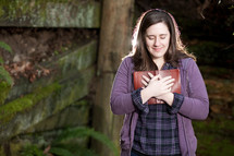 Woman standing outdoors praying with Bible held to her chest.