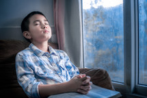 a boy sitting in a window praying with a Bible in his lap 