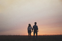 A man and woman hold hands in a field at dusk.