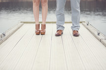 The feet and legs of a man and woman standing on a dock on a lake.