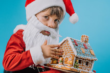 Cute little child in Santa Claus costume sitting with gingerbread house on blue background. Christmas celebration. Happy childhood, kid, lovely son. High quality photo